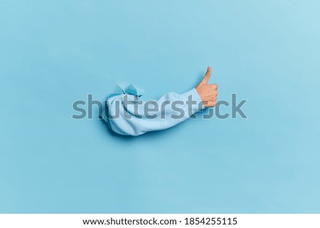 Human hand breaking through paper wall and showing thumb up as sign of approval or agreement isolated over blue studio background. Place for text information. Acceptance okay and liking concept