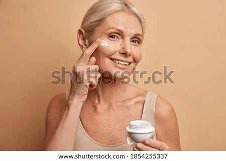 Smiling tender woman holds jar of cream with anti aging effect applies on face has healthy glowing skin looks pleasantly at camera undergoes beauty treatments isolated over brown background.