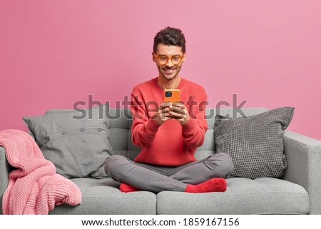 Cheerful technology addicted guy relaxes on comfortable couch holds modern mobile phone chats in social networks watches funny videos online spends weekend at home checks application or email box