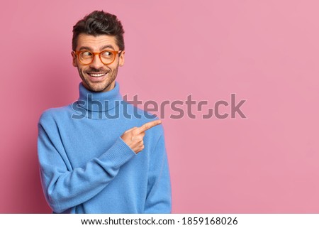 Use this copy space wisely. Glad handsome man in casual outfit points away on right advetises something smiles happily wears blue turtleneck isolated over pink background shows good offer to you