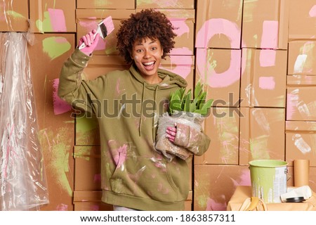 Joyful dark skinned woman painter renovates new house moves in apartment paints walls holds paintbrush and pot of green cactus improves room smiles broadly. Home decorating and improvement concept