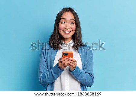 Beautiful Asian woman holds modern cellular glad to surf in social networks smiles positively dressed in casual jumper poses against blue background. Technology and online communication concept