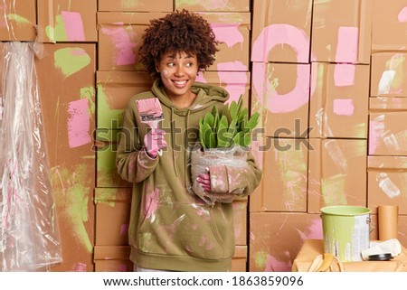 House improvement and renovation concept. Happy curly haired woman wears casual sweatshirt stands dirty holds paint brush potted cactus moves in new flat being busy with repair. Female painter