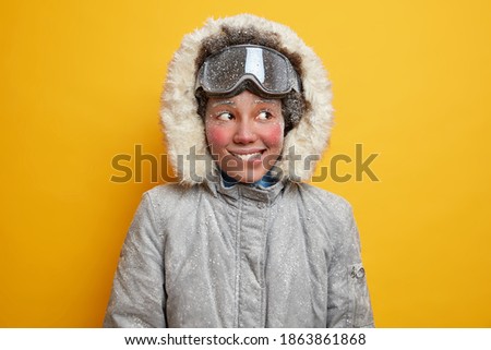 Portrait of cheerful frozen woman bites lips and looks away gladfully has adventure or winter expedition in tundra has hoarfrost face dresses for cold climate hiking in blizzard wears warm jacket