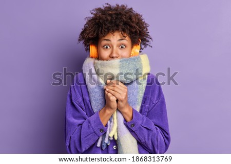 Afro American woman feels very cold during freezing weather wears purple jacket and warm scarf around neck wals on street during winter listens music via wireless headphones keeps hands together 商業照片 © 