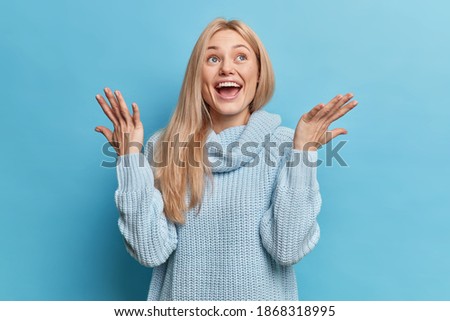 Horizontal shot of overjoyed blonde European woman raises hands and laughs positively sees something awesome wears knitted sweater poses against blue background. Happy emotions and reaction concept