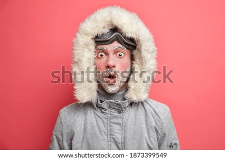 Stupefied emotional male tourist spends winter vacation in mountains enjoys active sport outdoors keeps mouth opened from great wonder covered with hoarfrost wears jacket and snowboarding goggles