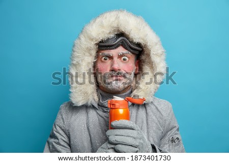 Puzzled frozen man in winter clothes tries to warm himself with hot beverage has red face and bear covered with blizzard spends much time outdoors during snowboarding. Frosty weather conditions