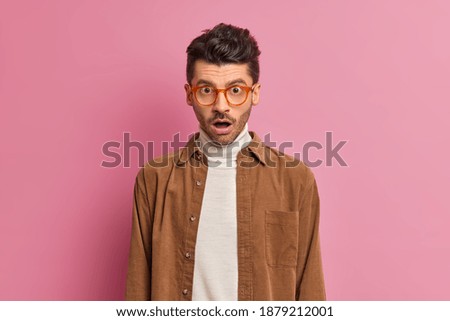 Speechless unshaven man looks in stupor drops jaw from astonishment cannot believe in shocking relevation wears optical glasses and brown shirt poses against pink background. Human emotions concept Foto d'archivio © 