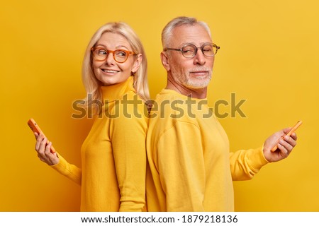 Studio shot of mature male and female models stand back to each other with modern gadgets in hands surf internet chat in social networks isolated over yellow background. Technology age concept