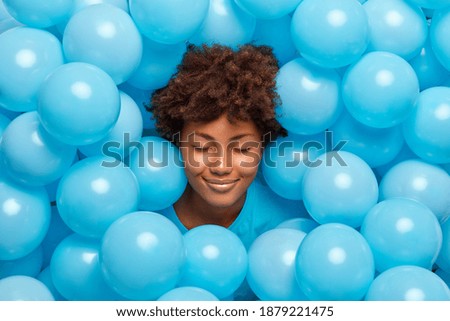 Pleased curly haired woman closes eyes surrounded by many blue inflated balloons has festive mood has fun on party feels very happy. Afro American birthday girl keeps head through decorated background