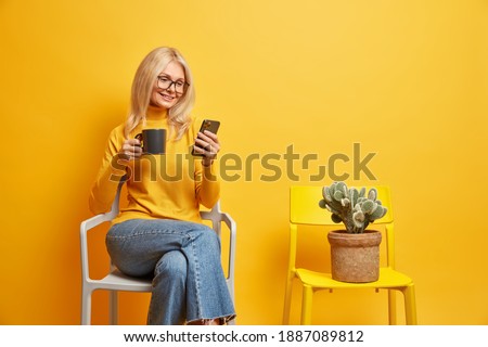Pleased middle aged woman works distantly from home has coffee break sits on comfortable chair surfs internet via mobie phone has glad expression dressed casually isolated over yellow background