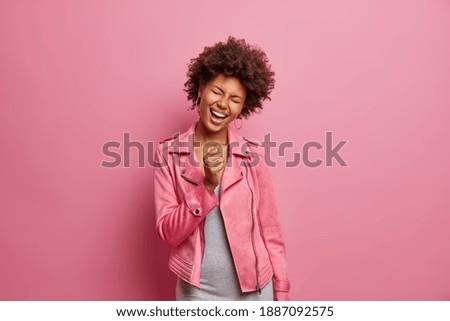 Glad young African American woman sings favorite song along keeps hand clenched in fist as if microphone has fun wears fashionable jacket isolated over pink background. Happy emotions concept