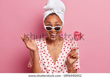 Positive good looking dark skinned female model holds delicious candy on stick wears wrapped bath towel sunglasses and domestic silk robe isolated over pink background. People and style concept