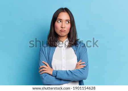 Portrait of displeased offended young Asian woman with dark hair keeps arms folded looks angrily aside doesnt agree with somebodys opinion wears neat clothes isolated over blue studio background