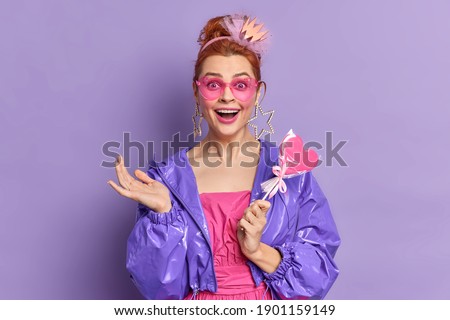 Retro fashion model dressed in nineties style has happy expression nostalgia holds delicious candy poses over vivid purple background. Fashion trends. Redhead girl in trendy sunglasses purple jacket