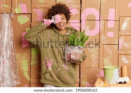 Cheerful curly Afro American woman covers eye with painting brush has fun while renovating new apartment holds cactus in pot poses against wall smeared with paints. House improvement concept