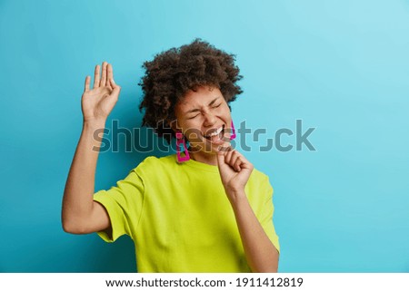 Positive African American woman sings song keeps hand as if microphone has fun has happy mood dressed in casual t shirt isolated on blue background. Carefree ethnic girl pretends to be in karaoke club