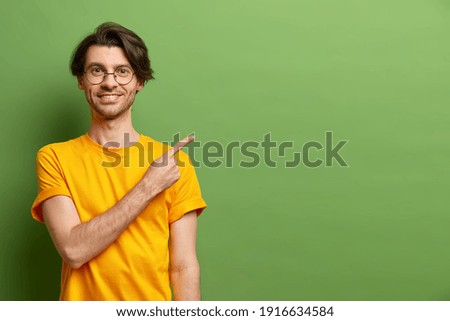 Cheerful Caucasian man points index finger at copy space suggests special offer dressed casually wears round glasses isolated over green studio background. Best choice. Place your advertisement there