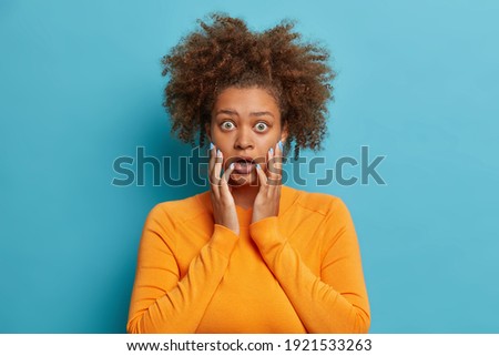 Studio shot of terrified curly haired teenager girl grabs face has eyes popped out stares in disbelief at camera wears casual orange sweater isolated over blue background. People reaction emotions