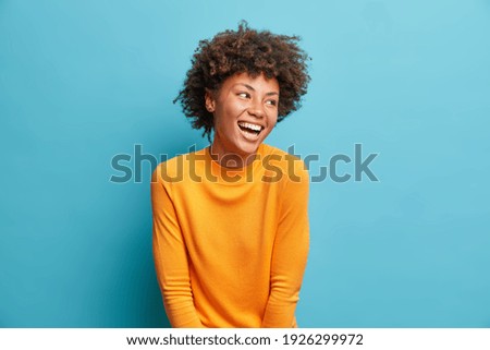 Happy emotions concept. Positive dark skinned beautiful young woman laughs poisitively looks aside with carefree face expression wears casual orange sweater isolated over blue studio background.
