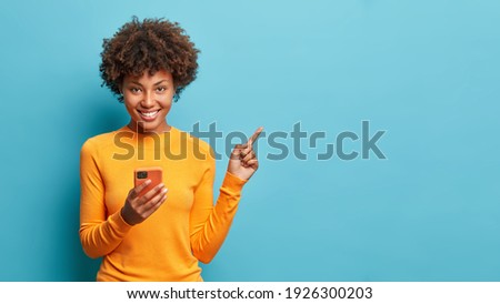 Foto horizontal de mulher afro-americana de pele escura feliz gosta de comunicação móvel e tecnologias modernas posa contra fundo azul aponta para o espaço livre para o seu conteúdo publicitário