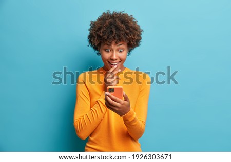 Happy surprised Afro American woman browses internet and networking in social media tests new application for smartphone wears casual jumper isolated over blue background. Mobile communication