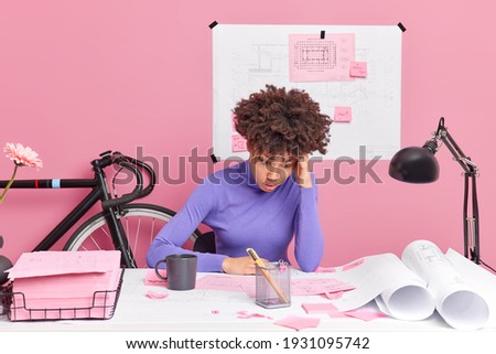 Puzzled woman architect improves graphics corrects mistakes busy working on archiectural project and having deadline poses at desktop has deadline surrounded with papers tries to solve problem