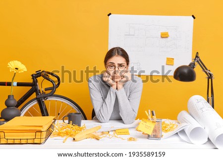 Sad bored professional woman architect makes building sketches and creates project leans hands under chin poses in coworking space feels tired during work day in office. Working process in workshop