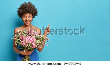 Happy African American woman with curly hair holds bouquet of flowers wears festive dress indicates at copy space shows advertisement or promotion enjoys spring and holidays isolated on blue wall