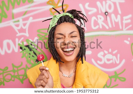 Trendy cheerful hipster girl smiles broadly holds lollipop has fun with teenagers of same age wears yellow vest poses against colorful graffiti wall. Gen z generation. Youth lifestyle concept Stok fotoğraf © 