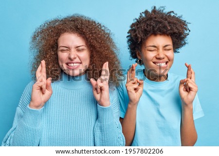 Studio shot of multiethnic women stand next ot each other cross finger for good luck anticipate positive news or result close eyes pose optimistic drssed casually isolated over blue background
