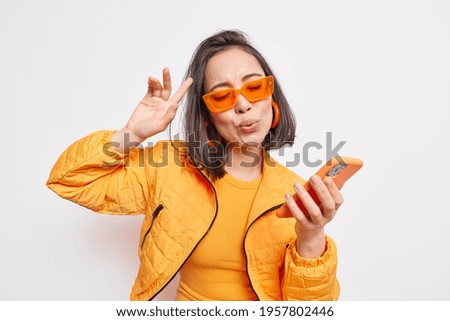 Cheerful brunette Asian woman dance to favorite song moves to rhythm of music holds modern smartphone wears trendy orange sunglasses stylish jacket expresses happiness and joy models indoor.