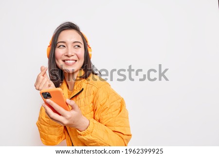 Studio shot of happy Asian millennial girl makes eastern like sign smiles gladfully looks away dressed in orange jacket uses smartphone and headphones for listening favorite music poses indoor