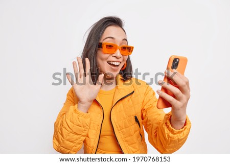 Positive stylish female blogger waves hi to followers in social networks wears orange jacket and sunglasses makes greeting gesture at smartphone isolated over white background connects to friends chat