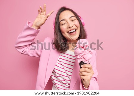 People street food lifestyle concept. Carefree happy Asian woman dances carefree raises arms being in good mood eats delicious ice cream closes eyes from pleasure sings favorite song uses headphones