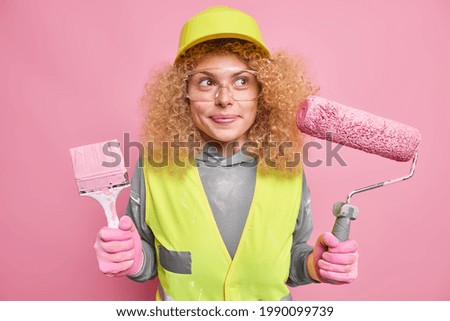 Repair service. Pleased thoughtful woman painter with curly fair hair holds brush and roller thinks about interior design of new dwelling does manual work dressed in uniform protective helmet