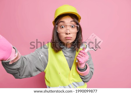 Shocked Asian woman builder makes selfie stands stunned indoor wears helmet uniform reflective vest paints walls of dwelling busy doing repair isolated over pink background. Remodeling redecorating