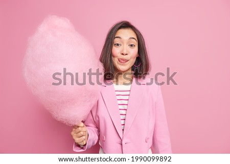 Good looking brunette Asian woman licks lips as holds appetizing sweet candy floss dressed in fashionable formal outfit isolated over pink background. Teenage girl with delicious cotton candy