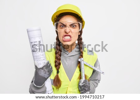 Irritated female builder clenches teeth angrily holds paper blueprint and tape measure going reconstruct dwelling wears safety helmet protective glasses uniform isolated over white background