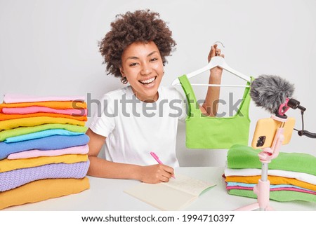 Smiling female fashion blogger demonstrates trendy clothes sells green top online makes notes about customer orders poses in front of camera at white table with colorful folded laundry near.