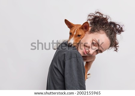 Sideways shot of adult girl carries puppy which embraces her around neck walkk together isolated over white background. Female dog lover poses with pet looks away likes animals. Care and love