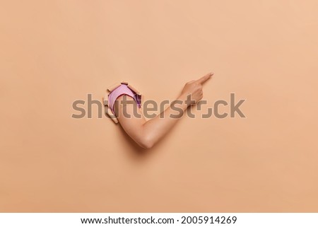 Thin nice girly arm goes through the hole in the paper wall and points out to something on the side. Wears beige band aid after the injection and pink tshirt. Isolated over beige background. 