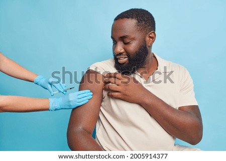 Dark skinned calm interested young man in a beige tshirt is frozen while getting the injection. A female nurse in turquoise gloves presses the skin before vaccination. Isolated over blue background.