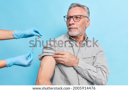 Retired bearded caucasian man in a grey shirt wears glasses, raises his sleeve for the nurse, prepares for the injection. An unrecognisable person in blue medical gloves removes the syringe cap. 
