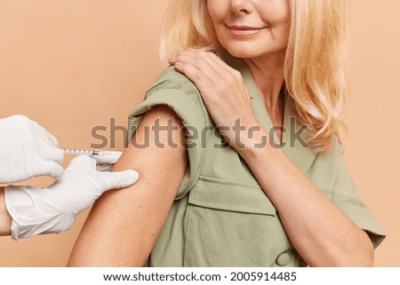 Unrecognizable satisfied caucasian light haired woman holds her shoulder, watches an injection, cares about her health and future. Hands in white medical gloves hold the shoulder for the injection. 