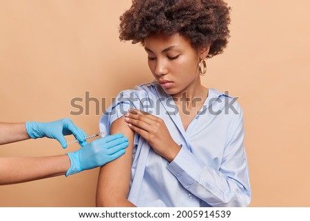 Pretty black lady is lost in her thoughts and worries about the pandemic, tries to stay calm and get the injection. Medical worker in blue gloves regains girl's composure and insert the syringe.