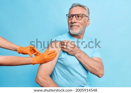 Bearded caucasian aged man in glasses is nervous about the injection, leans backward, afraid of vaccination. Kind unknown nurse in orange gloves helps calm the patient. Isolated over blue background.