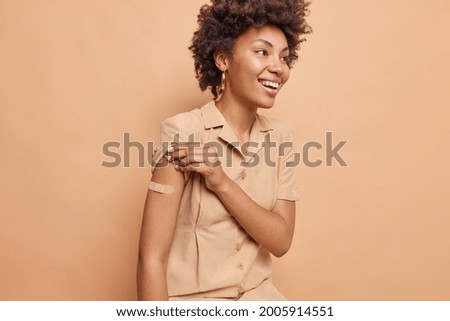 Young curly haired African American woman in a beige dress is happy about the first vaccination, wears band aid, raises sleeve and tells friends about the injection. Isolated over beige background.
