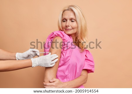 Beautiful thoughtful caucasian blond longhaired adult woman holds her elbow for the nurse to get an injection, wears a pink loose dress. Nurse holds the injection site, wears white medical gloves.